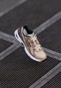 Baskets de course New Balance beiges et marron avec une semelle blanche reposant sur un sol en grille métallique.