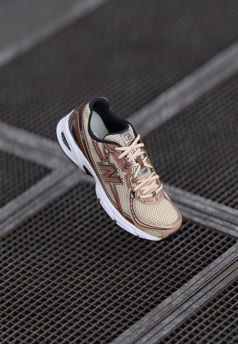 Baskets de course New Balance beiges et marron avec une semelle blanche reposant sur un sol en grille métallique.