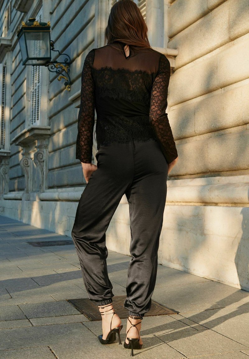 Schwarzer Jumpsuit mit langen Spitzenärmeln, am Handgelenk eng anliegend. Kombiniert mit schwarzen, schnürbaren High Heels. Glatter Stoff, eleganter Schnitt.