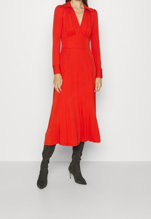 Femme portant une robe midi rouge vif à manches longues avec un décolleté en V profond et des bottes en daim noires à talons hauts, debout devant un fond uni.