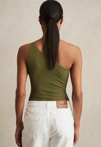 Top oliva verde a costine con una spalla e dettagli in pizzo, abbinato a pantaloni bianchi a vita alta con una etichetta in pelle marrone con marchio.