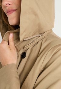 Chaqueta con capucha beige con cierre de cremallera, detalle de botón grande y textura de tela suave. Vista en primer plano mostrando una mano ajustando el cuello.