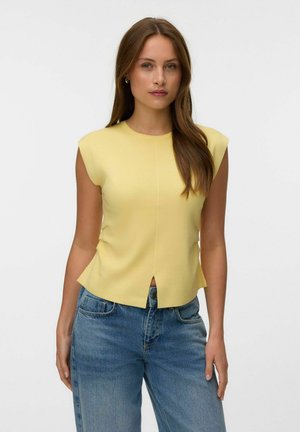 Jeune femme aux longs cheveux bruns portant un haut jaune sans manches et un jean bleu, debout devant un fond clair uni.
