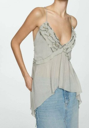 Kobieta w przezroczystym, jasnoszarym, bezrękawnikowym topie z falbankami i cekinami, w połączeniu z jasnoniebieskimi dżinsami z wysokim stanem.