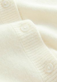 Tissu en tricot couleur crème avec une texture douce, présentant un col côtelé et trois boutons en plastique blanc le long du bord.