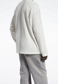 Osoba ubrana w długi rękaw beżowy top w prążki i luźne szare spodnie, pokazana od tyłu na tle białego tła.