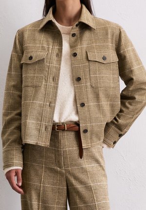 Kobieta w beżowej, kratkowanej, zapinanej na guziki kurtce i dopasowanych spodniach z brązowym paskiem, na kremowym swetrze, z ręką na biodrze.