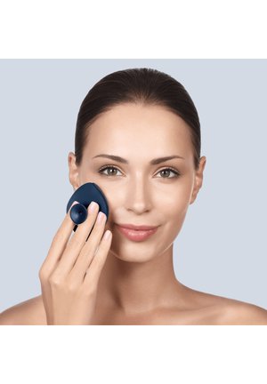 Woman with pulled-back hair uses a dark blue silicone facial cleansing brush on her cheek against a light gray background.