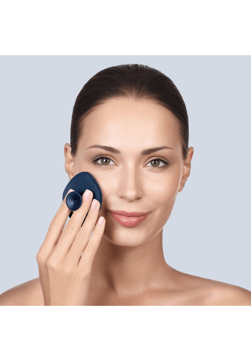 Donna con capelli raccolti usa una spazzola per la pulizia del viso in silicone blu scuro sulla guancia su sfondo grigio chiaro.