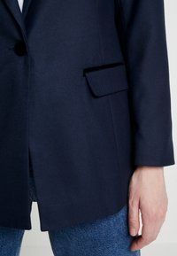 Blazer bleu marine avec bouton noir, poche à rabat, porté sur un jean bleu, montrant la manche et la main d'une personne.