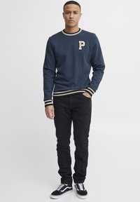 Marineblauwe sweatshirt met een beige "P" embleem, ribgebreide kraag en zoom met romige en zwarte strepen, gecombineerd met zwarte broek en zwarte sneakers.