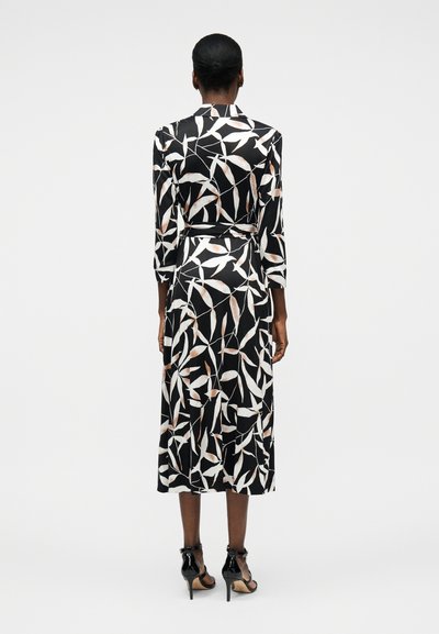 Γυναίκα που στέκεται με πλάτη, φοράει μαύρο μίντι φόρεμα με λευκό και μπεζ σχέδιο φύλλων, μακριά μανίκια, ζώνη και μαύρα σανδάλια με ψηλό τακούνι.
