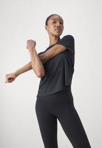 Czarna koszulka z krótkim rękawem o luźnym kroju, połączona z czarnymi prążkowanymi legginsami. Modelka rozciąga się, unosząc jeden ręka. Gładkie tło.