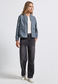 Veste bomber bleu clair avec des poignets élastiques et deux poches avant, associée à un jean gris foncé et des baskets blanches. Tissu lisse et brillant.