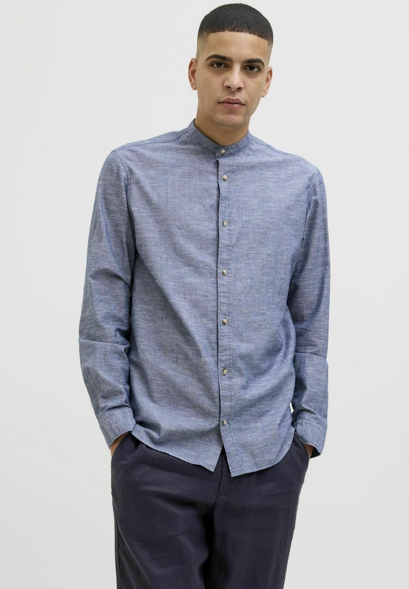Camicia button-up azzurro chiaro con colletto alto, tessuto dalla texture visibile e bottoni in vista sul davanti, abbinata a pantaloni scuri.
