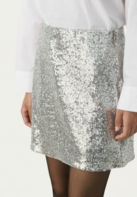 Zilveren pailletten mini-jurk met een zijrits, een gladde textuur en een glanzende afwerking. Gepaard met een wit shirt met lange mouwen en zwarte panty.