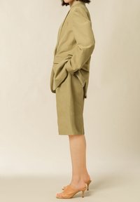 Blazer su misura verde oliva chiaro e pantaloni corti abbinati fino al ginocchio, abbinati a décolleté beige. Il tessuto appare liscio con minima texture.