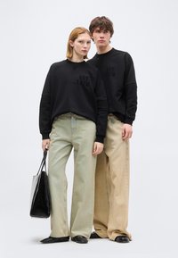 Δύο μοντέλα φορούν ταιριαστά μαύρα φούτερ με την επιγραφή "EST 1978," φαρδιά μπεζ τζιν και μαύρα παπούτσια· το ένα κρατά μια μεγάλη μαύρη τσάντα tote.