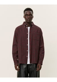 Camicia a quadri rossa e verde con colletto, maniche lunghe, caratterizzata da una tasca frontale e una vestibilità comoda. Indossata sopra una maglietta bianca.