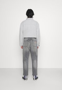 Chaqueta de mezclilla gris con detalles de botones, combinada con jeans grises que tienen un logo en el bolsillo trasero. Zapatillas negras y blancas visibles.