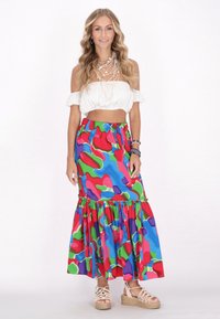Fustă maxi colorată, cu modele și margine ondulată în albastru, roșu, verde și mov; asortată cu un top alb scurt cu umerii la vedere și espadrile.