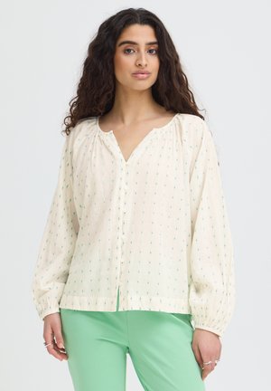 Žena s dlhými kučeravými vlasmi, ktorá má na sebe krémovú blúzku s malými zelenými bodkami a zelené nohavice, stojí pred jednofarebným pozadím.