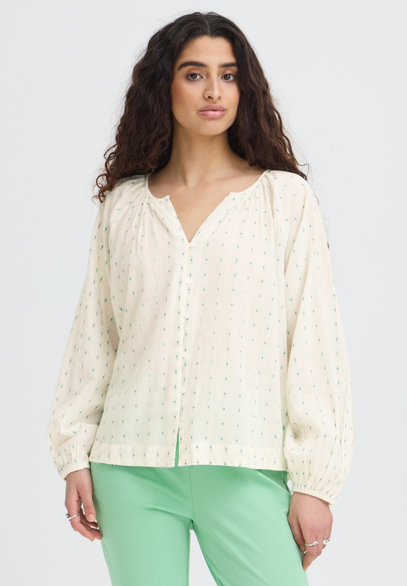 Femme aux longs cheveux bouclés portant un chemisier crème à petits pois verts et un pantalon vert, debout sur un fond uni.