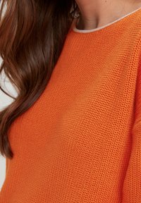 Suéter naranja con un patrón de punto texturizado, que presenta un cuello de color claro en contraste. Primer plano, destacando los detalles de material y color.