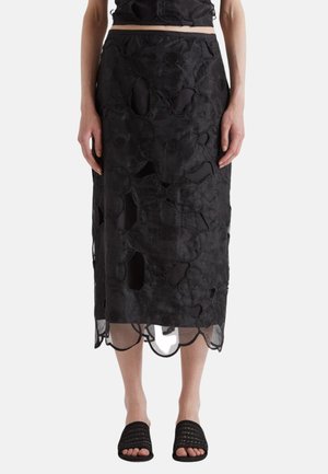 Kvinde iført sort blomsterbroderet midi-nederdel og matchende cropped top med sorte åbne vævede sandaler, stående mod en ensfarvet baggrund.