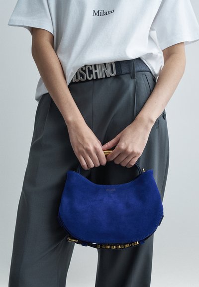 Osoba držící modrou semišovou kabelku se zlatou rukojetí, oblečená v šedých kalhotách a bílém tričku s nápisem "Milano", a s pásem MOSCHINO.