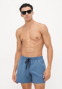 Blå svømmetights med elastisk livlinje, sort snor og en lett, glatt tekstur. Båret av en modell med solbriller.
