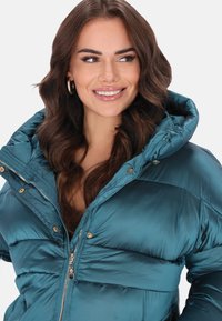 Turkusowa pikowana kurtka puchowa z błyszczącym wykończeniem, zapięciem na zamek i kapturem. Posiada pikowane sekcje oraz złote ozdoby. Lekka i ocieplana.