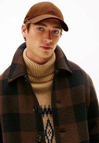 Giovane uomo che indossa un cappello di velluto a coste marrone, un maglione a collo alto beige con motivo e un cappotto a scacchi nero e marrone scuro.