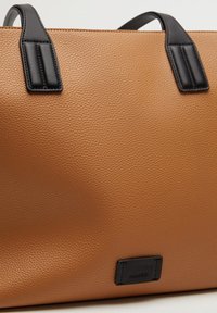 Duża beżowa torba na ramię wykonana z teksturowanej sztucznej skóry, z czarnymi uchwytami i małą czarną metką z logo na dole.