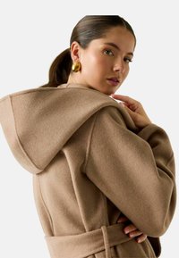 Cappotto di lana marrone con cappuccio ampio, vita con cintura e tessuto morbido, caratterizzato da una silhouette rilassata e dettagli minimali.