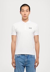 Hvit polo skjorte med klassisk krage og korte ermer. Har en liten svart EA7-logo på venstre bryst. Stoffet ser glatt ut.