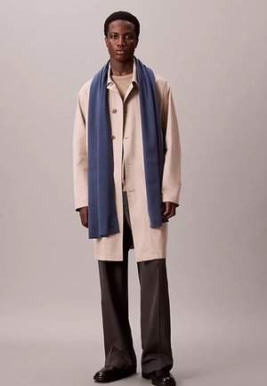 Mann trägt einen beigen Trenchcoat über einem hellen Pullover, weite dunkle Hose, schwarze Schuhe und einen langen blauen Schal vor einfarbigem Hintergrund.