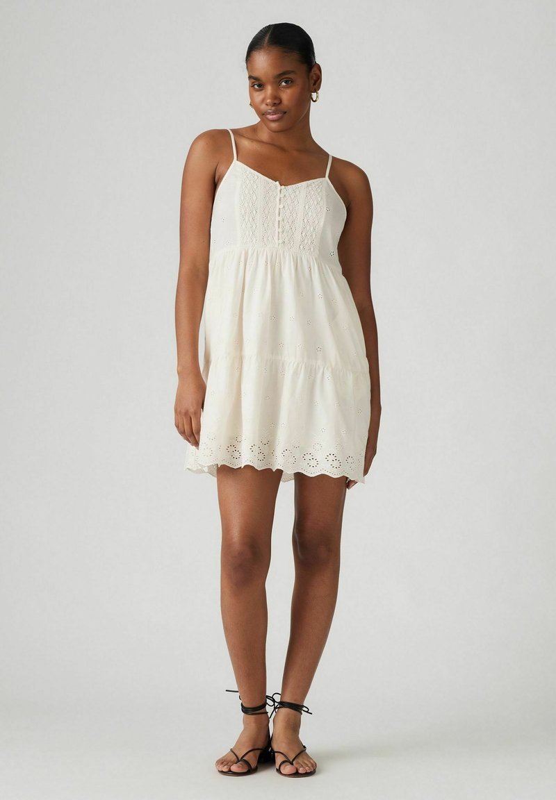 Vestido blanco de algodón con acentos de encaje, un corsé ajustado y una parte inferior acampanada. Las tirantes de espagueti y un dobladillo con forma de concha completan el diseño.