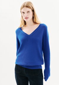 Maglione a maniche lunghe a V in tessuto lavorato di un blu brillante; presenta una vestibilità comoda e una texture morbida. Indossato con jeans scuri.