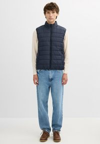 Chaleco sin mangas acolchado azul marino con cuello alto, combinado con una camisa de manga larga ligera, vaqueros sueltos de color azul claro y zapatos marrones con cordones.