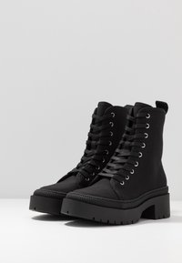 Bottes cheville noires en tissu avec une finition texturée. Elles présentent une semelle épaisse, un talon surélevé et des œillets en métal pour les lacets.