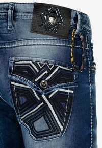 Jean en denim avec une poche noire texturée ornée de broderies géométriques, une étiquette en cuir noir avec un logo, et des coutures blanches contrastantes.