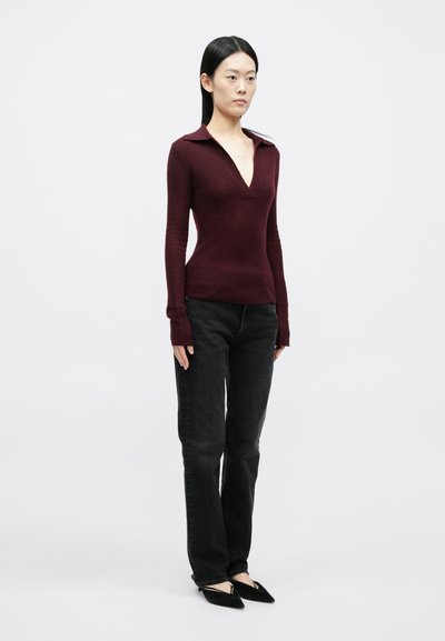 Γυναίκα στέκεται φορώντας εφαρμοστή βυσσινί μπλούζα με μακριά μανίκια και βαθύ V-λαιμόκοψη, μαζί με μαύρο τζιν με ίσια γραμμή και μαύρα μυτερά παπούτσια.