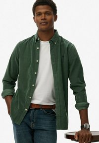 Donkergroen button-up shirt van stof, gedragen over een wit T-shirt. Gecombineerd met blauwe jeans en een bruine riem, met een horloge om de pols.