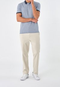 Man i beige byxor, grå polo och vita sneakers står med en arm korsad och handen på hakan mot en vit bakgrund.