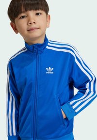 Blå zip-up träningsoveralljacka med vita tre-randiga detaljer på ärmarna, ribbade muddar och krage, med en Adidas-logotyp på bröstet.