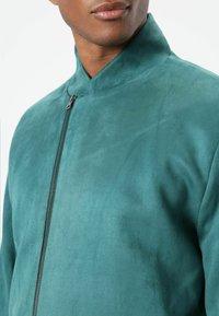 Groeneblauwe jas met een hoge kraag, gemaakt van een gestructureerde, zachte stof. Een rits aan de voorkant benadrukt het strakke, minimalistische ontwerp.