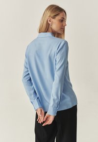 Cămașă albastru deschis, cu mânecă lungă, fabricată dintr-o țesătură netedă, cu guler și manșete butonate. Spatele are un croi drept, cu un design minimalist.