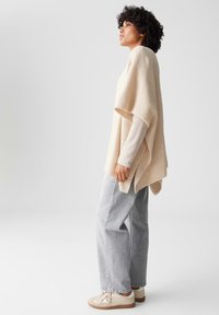 Beiger gerippter Poncho, lockerer Schnitt, kombiniert mit hellgrauen weit geschnittenen Jeans und cremefarbenen Sneakers mit beigen Akzenten.