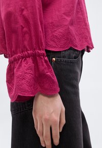 Φούξια μπλούζα με κεντημένα μανίκια με κυματιστό τελείωμα, κατασκευασμένη από ελαφρύ ύφασμα, συνδυασμένη με σκούρο τζιν. Διαθέτει λεπτομέρεια μανικιού με σούρες.
